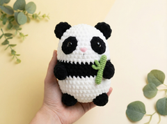 A hand holds a crocheted panda plush toy holding a green bamboo shoot, set against a warm beige background with decorative green leaves.