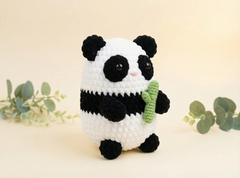 A crocheted panda toy holding a green bamboo shoot, set against a warm cream background with decorative eucalyptus sprigs.