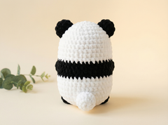 A crocheted panda toy shown from the back, featuring a white body with black ears, black stripes, and black feet, set against a warm beige background with a small green plant sprig on the left.
