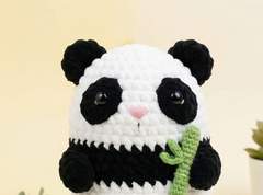 A crocheted panda plush toy holding a green bamboo shoot, featuring black and white yarn, a pink nose, and glossy black eyes, set against a soft beige background.
