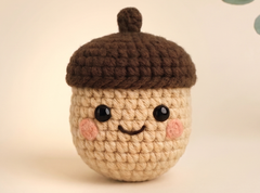 A cute crocheted acorn toy with a smiling face, featuring a beige body, a brown cap, and pink cheeks, set against a soft beige background.