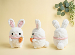 Three white crocheted bunny figures shown from front, side, and back views on a soft beige background, each holding a small orange carrot and wearing a pink band around the neck.