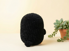 A black crocheted plush toy with a rounded, egg-shaped body and small tail, shown from the back on a light beige surface with a potted succulent in the background.