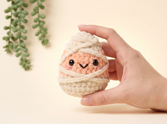 A hand holding a cute, amigurumi-style crocheted egg character wrapped in cream yarn, with a peach-colored face and embroidered black eyes and smile, against a warm beige background with a green plant in the corner.
