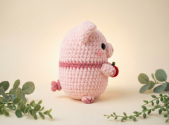 A pink crocheted plush pig toy holding a small red strawberry, photographed on a warm cream background with green eucalyptus sprigs around it.
