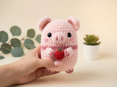 A hand holds a small crocheted pink pig toy with black eyes and a red strawberry accessory, set against a warm beige background with blurred green leaves and a small potted succulent.