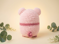 A pink crocheted plush toy shaped like a pig, shown from the back on a warm cream background with decorative green leaves around it.