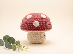 A crocheted mushroom toy with a dusty red cap, white spots, and a beige stem, styled with a green eucalyptus sprig on a warm cream background.
