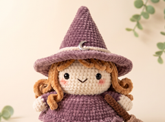 A crocheted doll with a pointed purple hat, cream-colored face and sleeves, and brown curly hair, shown against a soft beige background with subtle green leaf accents.