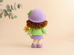 Amigurumi doll with curly brown hair, lilac dress and shoes, standing in a pastel nursery scene.