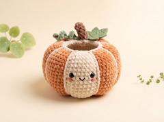 A cute crocheted pumpkin planter with a smiling face, featuring orange and cream stripes, green leaves, and a brown stem, set against a warm beige background with small green plant accents.