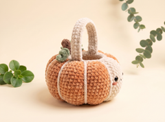 A crocheted pumpkin-shaped handbag with a cute face, featuring a warm orange body, cream handle and lower section, and small green stem accents, styled on a beige background with eucalyptus leaves.