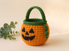A crocheted pumpkin-shaped candy bucket with a green handle and stem, featuring a black embroidered jack-o'-lantern face, set against a warm beige background with a decorative eucalyptus sprig.