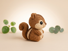 A small crocheted squirrel toy in brown and cream, posed on a warm beige background with soft green leaves blurred in the background.