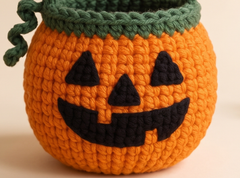 A crocheted pumpkin-shaped bucket in bright orange yarn with a dark green top trim and black embroidered jack-o'-lantern face details, shown on a plain light beige background.