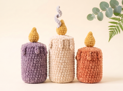 Three crocheted candle-shaped items in purple, cream, and terracotta orange, each with a small flame on top, arranged against a warm beige background with eucalyptus leaves in the corner.