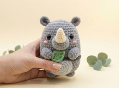 A small crocheted rhinoceros toy being held in a person's hand against a soft cream background with a few green leaves as props.