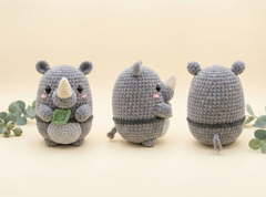 Three views of a crocheted gray rhinoceros plush toy on a warm beige background with decorative green leaves. The left view shows the front with a white horn and a green leaf, the center view shows the side, and the right view shows the back.