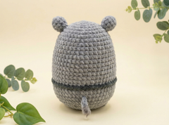 A gray crocheted plush toy shaped like a rounded character with two small ears on top and a short tail at the bottom, photographed on a soft cream background with decorative green leaves around it.