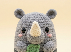 A crocheted rhinoceros toy with a gray body, cream-colored horn, black eyes, pink cheeks, and a green leaf in its front, set against a soft beige background.
