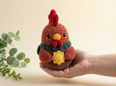 A crocheted amigurumi chicken doll held in a person's hand against a warm beige background, with a small green plant visible on the left.