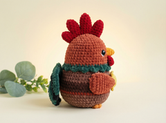 A crocheted toy rooster in warm brown and orange tones with a red comb and wattle, a dark green tail, and a yellow beak, photographed on a soft cream background with a small green leafy sprig at the side.