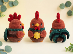 Three crocheted rooster amigurumi toys are displayed on a light cream background with decorative eucalyptus leaves. The center rooster is shown holding a yellow flower, while the left and right roosters show the front and back views respectively.