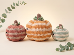 Three crocheted plush pumpkins in autumn colors, including a large striped orange and cream pumpkin in the center, flanked by two smaller striped pink and cream and mint and cream pumpkins, styled with eucalyptus branches on a warm cream background.