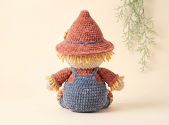 A crocheted scarecrow doll with a terracotta hat, yellow straw hair, a rust-orange shirt, and blue overalls, photographed against a warm cream background with a soft green leafy branch in the upper right.