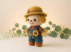A crocheted scarecrow doll stands centered against a warm beige background with soft green eucalyptus branches behind it. The doll wears a yellow straw hat, blue overalls, and an orange-brown shirt, with a yellow sunflower at its waist.