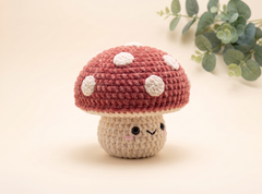 A crocheted mushroom figurine with a dusty rose red cap featuring white spots and a beige stem with a cute embroidered face, set against a warm cream background with eucalyptus leaves in the corner.