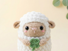 A crocheted plush sheep toy with fluffy white wool, a beige face and ears, pink embroidered cheeks, and a green four-leaf clover held in its front hooves, set against a soft beige background with blurred green leaves.