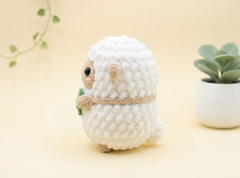 A small crocheted plush sheep figurine in white with beige ears, eyes, and hooves, holding a tiny green leaf, set against a soft beige background with a small potted succulent on the right.