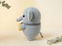 A crocheted plush elephant toy in light blue-gray, holding a small yellow honey pot, set against a warm cream background with decorative eucalyptus leaves.