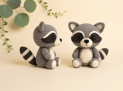 Two crocheted raccoon toys in gray, black, and cream are displayed on a warm beige background with decorative green leaves in the top left corner.