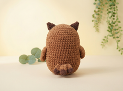 A brown crocheted plush toy shaped like a small animal or creature, shown from the back on a light cream surface with soft green leaves and hanging vines in the background.