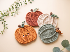 Four crocheted pumpkin-shaped coasters in orange, rust, cream, and sage green, arranged on a light beige background with decorative green leaves.