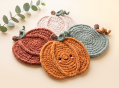 Four crocheted pumpkin-shaped coasters arranged on a light cream background, with a small green eucalyptus sprig in the top left corner. The front coaster is orange with embroidered facial features, layered over a dusty rose coaster, a mint green coaster, and a cream coaster.