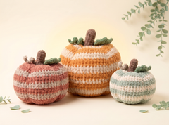 Three crocheted decorative pumpkins in muted fall colors, arranged on a warm cream background with soft eucalyptus leaves. The pumpkins have striped patterns in pink, cream, orange, and mint green, with brown stems and green leaves.