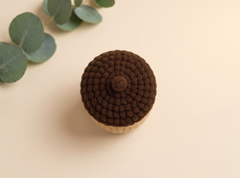 A crocheted acorn-shaped item in dark brown and beige, photographed from above on a warm cream background with eucalyptus leaves in the top-left corner.