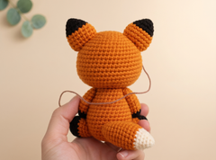 A hand holding a crocheted fox toy from behind, showing its orange body, black ears and paws, and white-tipped tail against a soft beige background.