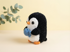 A crocheted penguin toy holding a blue fish, standing on a light beige surface against a pale yellow background with a soft-focus eucalyptus branch in the upper left.