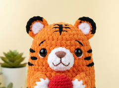 A close-up of a crocheted tiger plush toy with orange yarn, black stripes, a white snout and paws, and a red ball in its paws, set against a soft beige background with a small potted succulent in the corner.