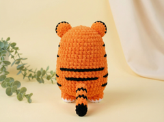 A crocheted orange tiger toy shown from the back, featuring black stripes, small ears, and a tail, set against a warm cream background with a green plant on the left.