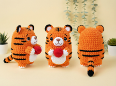 Three crocheted tiger plush toys are displayed on a light beige surface against a soft beige background with hanging greenery and small potted plants. The center toy faces forward holding a red heart, while the left toy faces left holding a red heart and the right toy faces away showing the back.