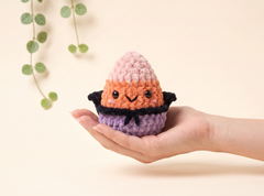 A small crocheted character egg with a pink and orange top, black collar, and purple base, held in a person's hand against a warm beige background with soft green leaves in the corner.
