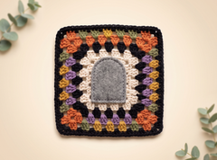A square crocheted textile piece with a central gray arch-shaped patch, surrounded by concentric rings of white, black, purple, orange, and olive green crochet motifs, photographed on a warm cream background with eucalyptus leaves in the corners.