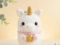 A cute crocheted unicorn plush toy with a white body, golden horn, pink collar, and star detail, set against a soft beige background with decorative eucalyptus leaves.