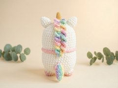 A crocheted unicorn plush toy shown from the back, featuring a white body, pastel rainbow mane and tail, pink horn, and small green eucalyptus leaves on a soft beige background.