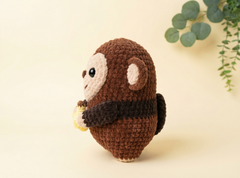 A crocheted monkey toy in brown and cream, holding a small yellow object, photographed on a warm cream background with a soft green plant leaf in the upper right corner.